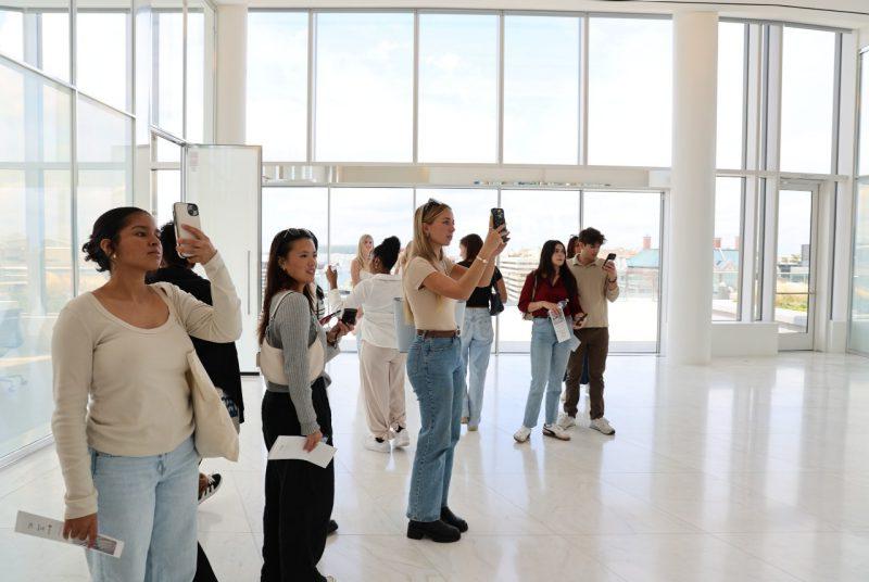 A group of students, some of whom are taking photos with cell phones, stands inside the interior of a building in front of a wall of windows